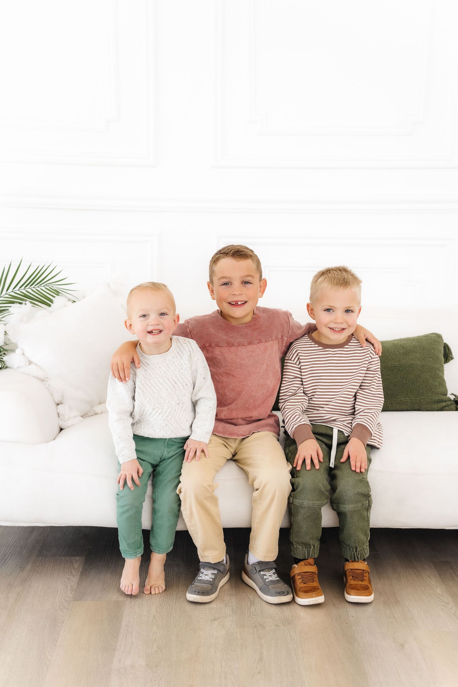Three young children sit together on a white couch wearing casual clothing in coordinated neutral tones. Best Family Photographer in Utah Fa