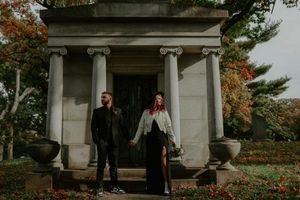 A couple poses together on the steps of a grand stone building with decorative columns and urns.