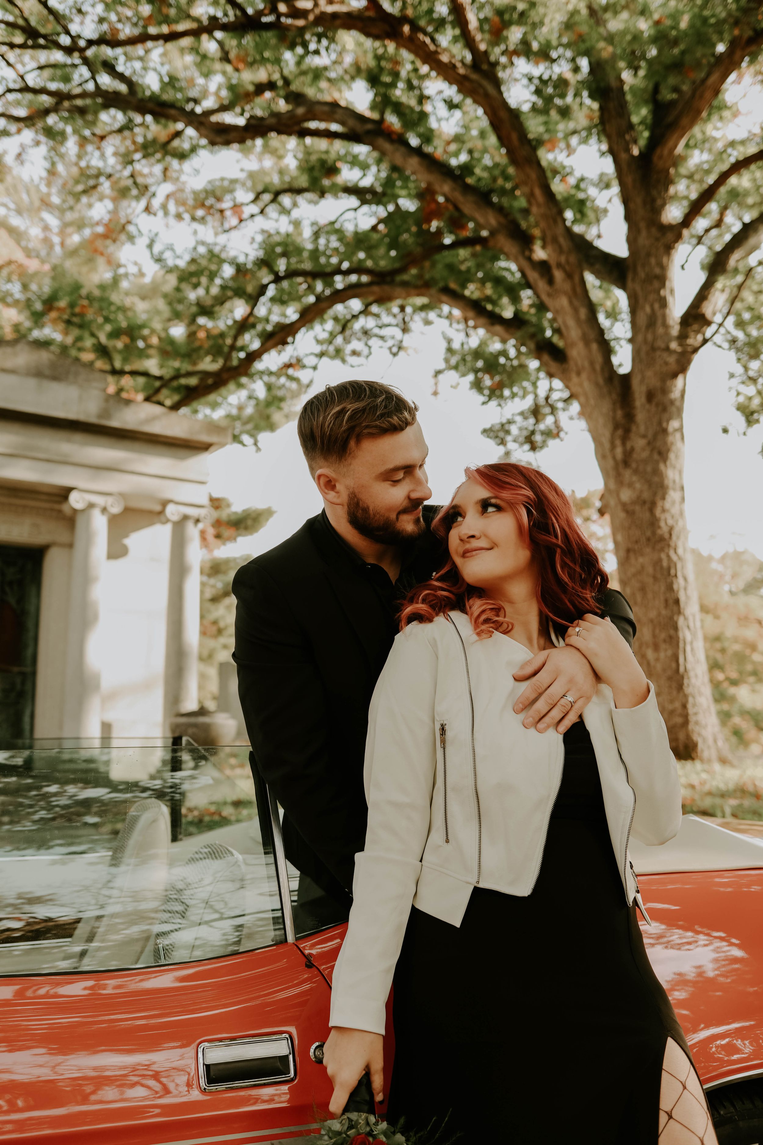 Couple sharing romantic moments next to a vintage red car under sprawling trees.