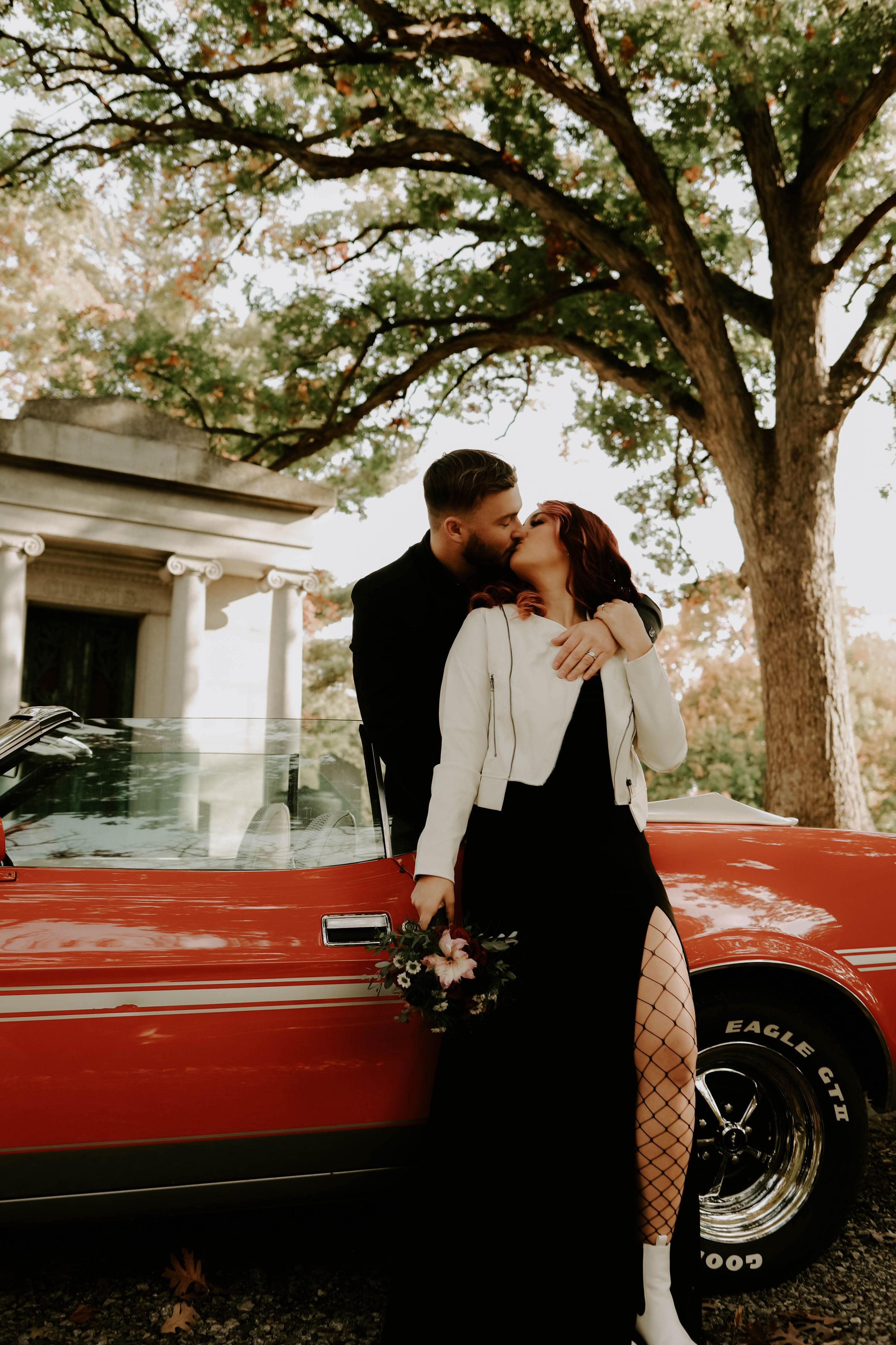 Stylish couple posing against a classic red car in a scenic outdoor setting.