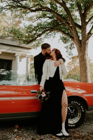A couple poses intimately next to a classic red muscle car under tall trees.