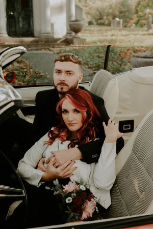 Couple in vintage attire pose romantically in a classic car with autumn scenery.