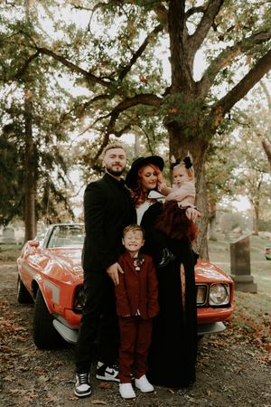 Family poses together near a vintage red car in an autumn cemetery setting.