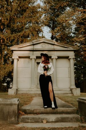 A figure in black and white stands on stone mausoleum steps surrounded by autumn trees in a moody cemetery setting.