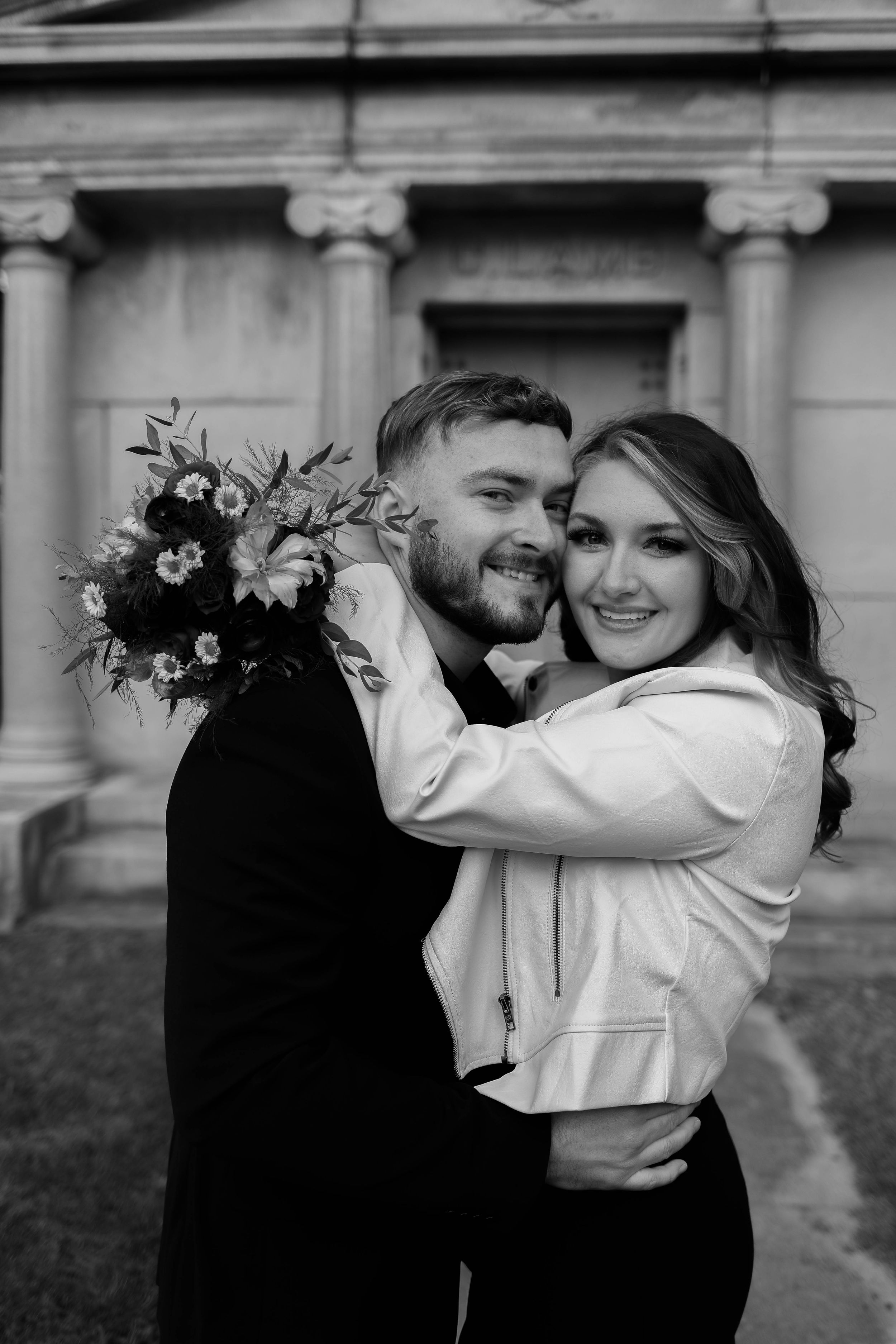 A couple embraces while holding a dark floral bouquet against classical architecture.