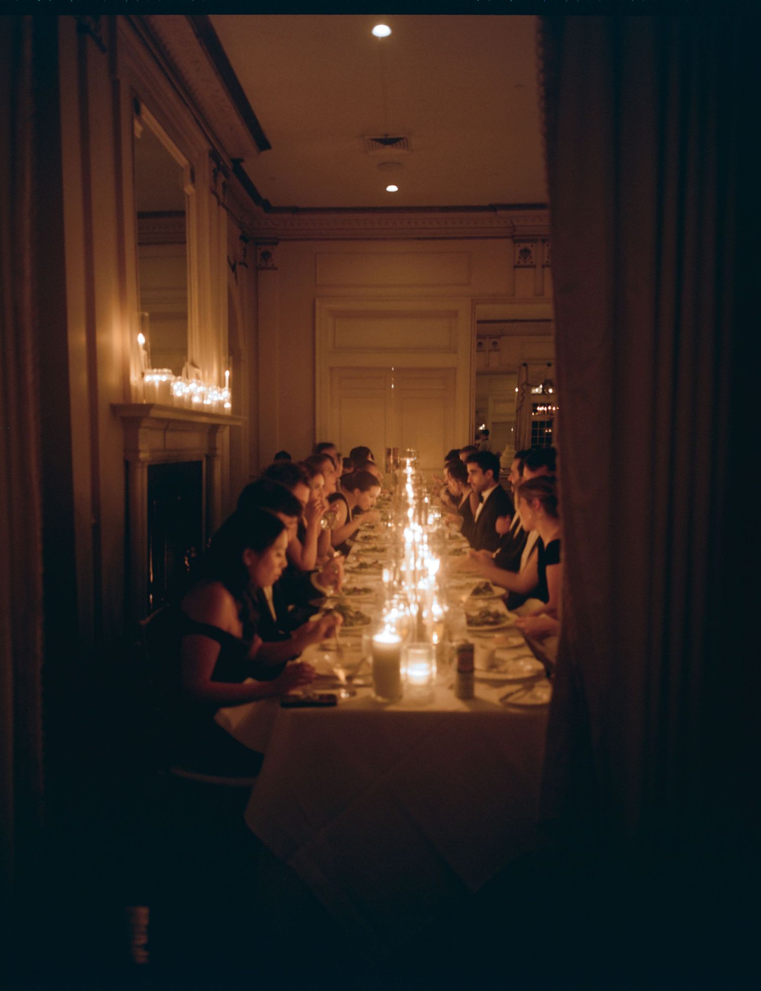 People gather around a long table illuminated by candlelight for an intimate dinner in a dimly lit elegant room.
