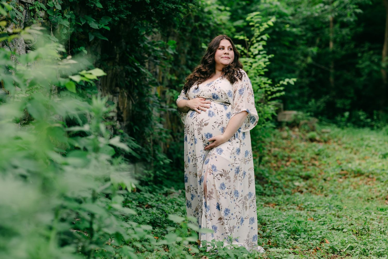 Someone in a floral maternity dress poses in a lush green forest setting during a maternity shoot at Southford Falls, CT