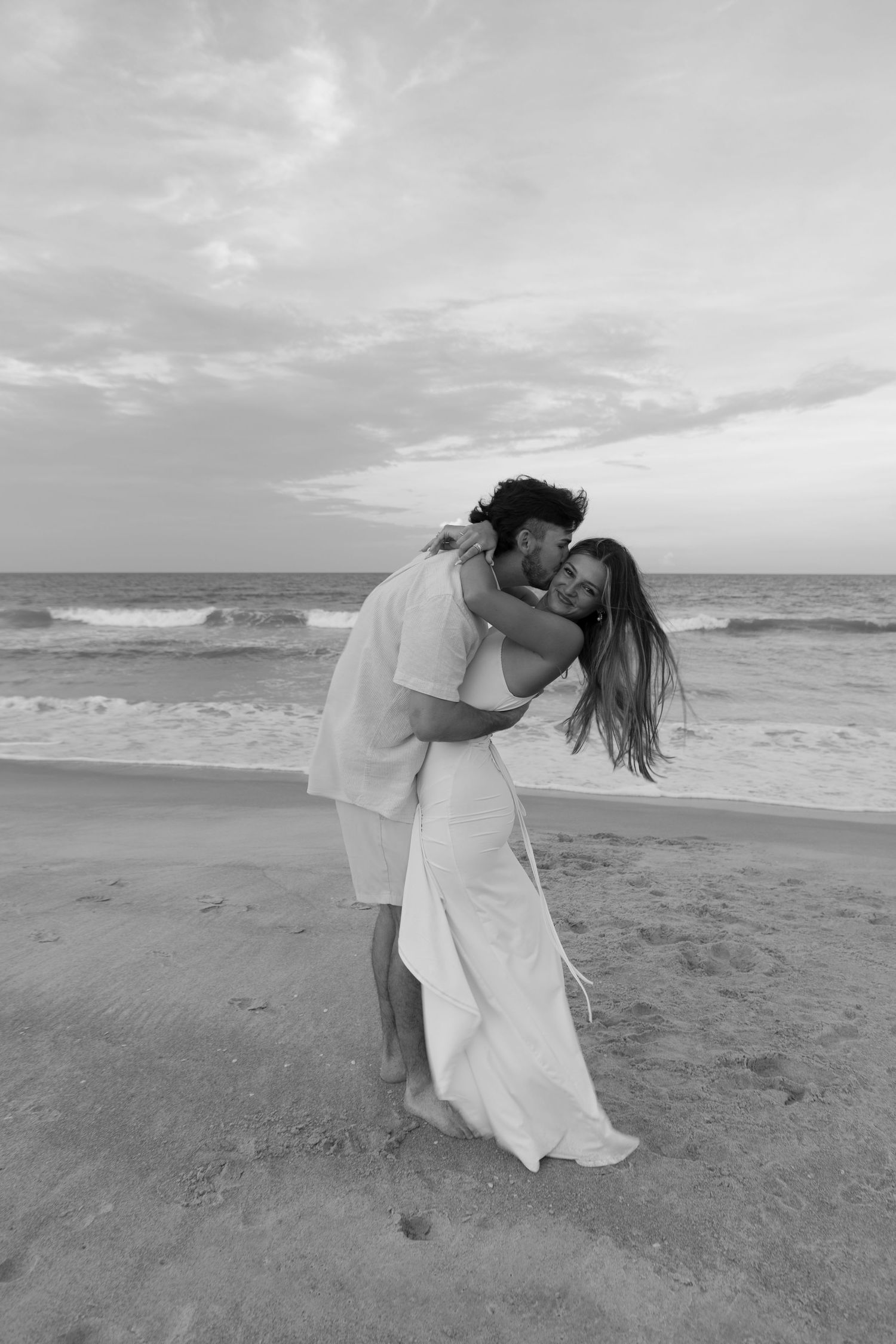 A couple shares a romantic kiss on a breezy beach at sunset in a stunning black and white photograph.