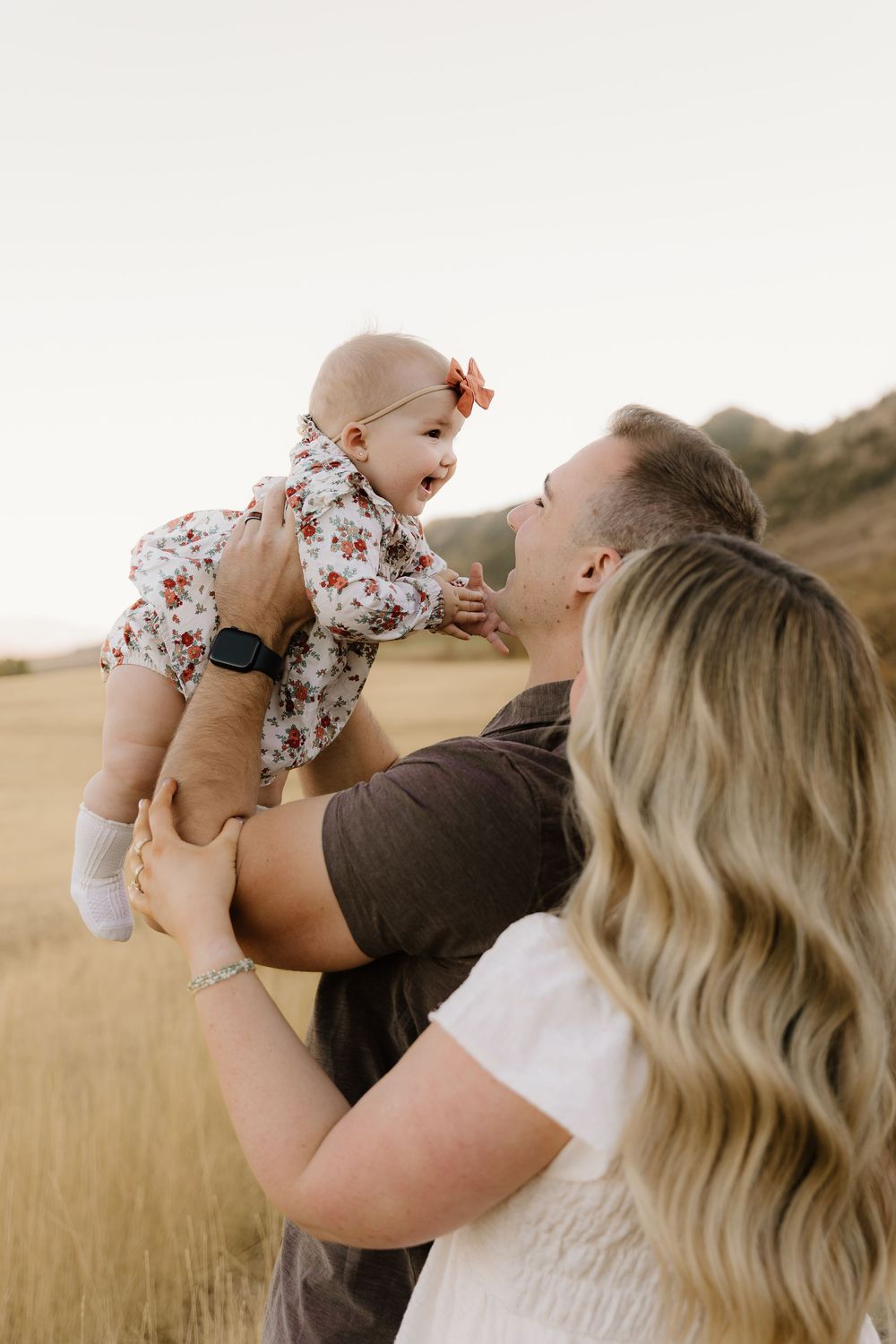 A joyful family moment captured in a golden field at sunset as they lift their baby in a floral romper.