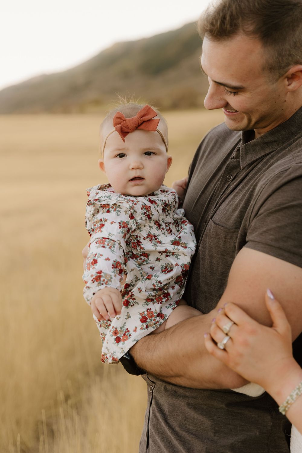 Parent holds baby wearing floral top in golden wheat field at sunset.