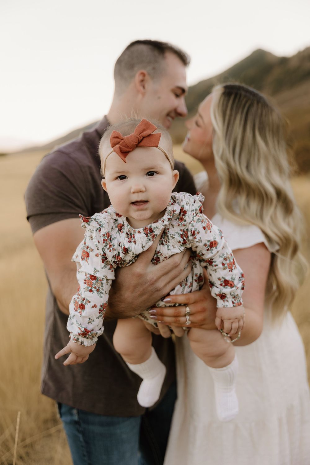Family portrait in golden field shows couple embracing while holding baby wearing floral outfit and orange bow.