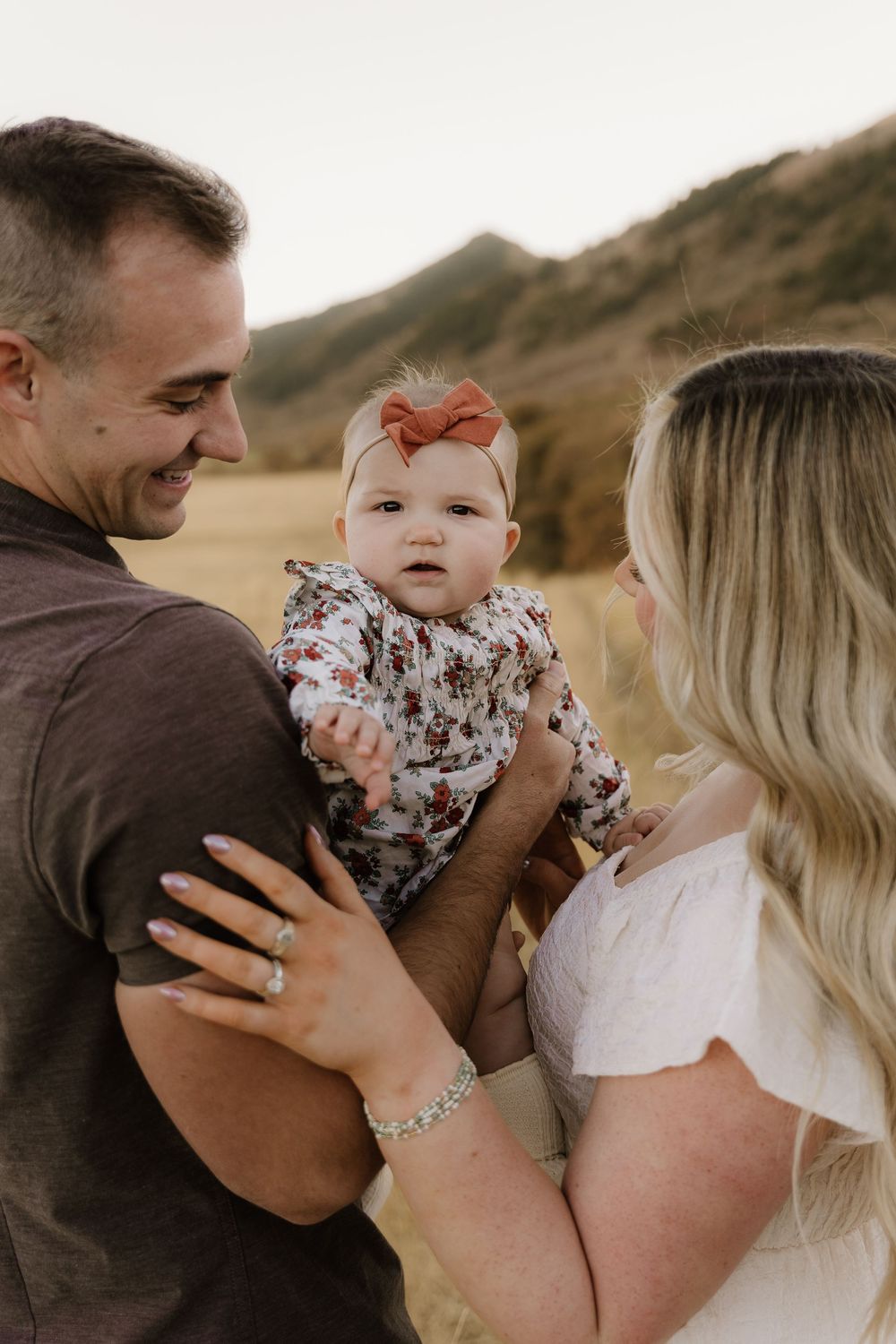 A joyful family moment outdoors with a baby wearing a floral outfit and orange bow against mountain backdrop.