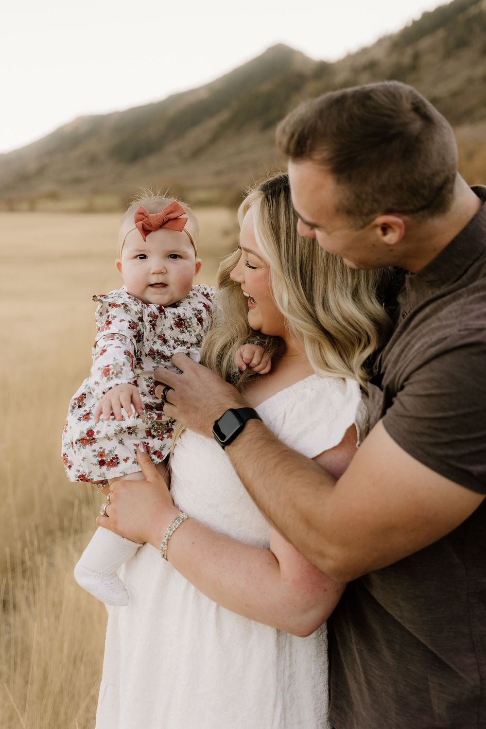 A loving family moment captured in a sun-lit field with mountains and tall grass in the background.