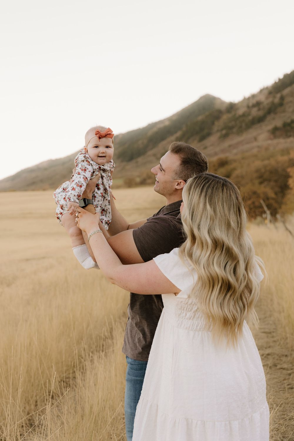 A couple in white and brown clothing lift their baby in a scenic golden field with mountains in the background at sunset.
