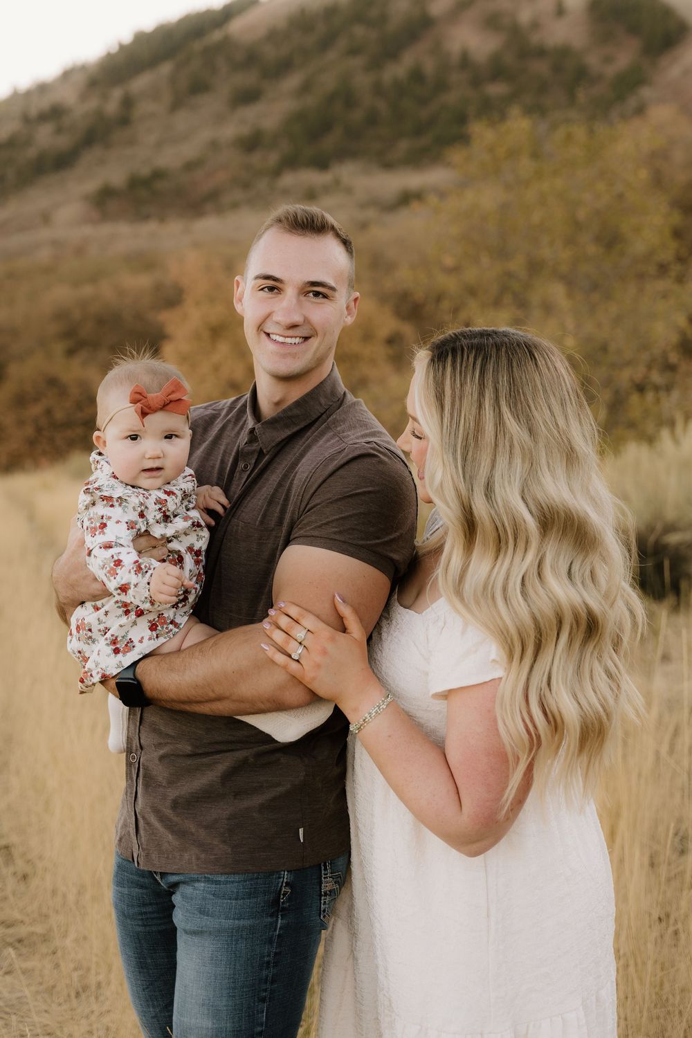 A family of three poses together in golden grass with mountains behind them at sunset.