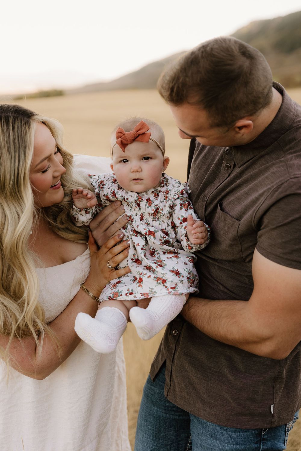 A loving family moment in nature as parents hold their baby wearing a floral dress and red headband in warm lighting.