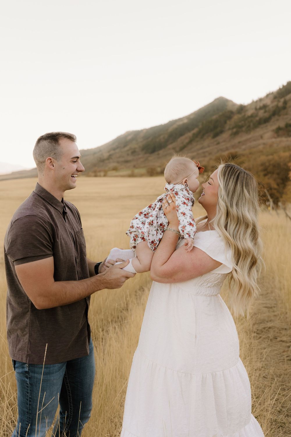 Candid family portrait in golden field shows joyful interactions between parents and baby against mountain backdrop.