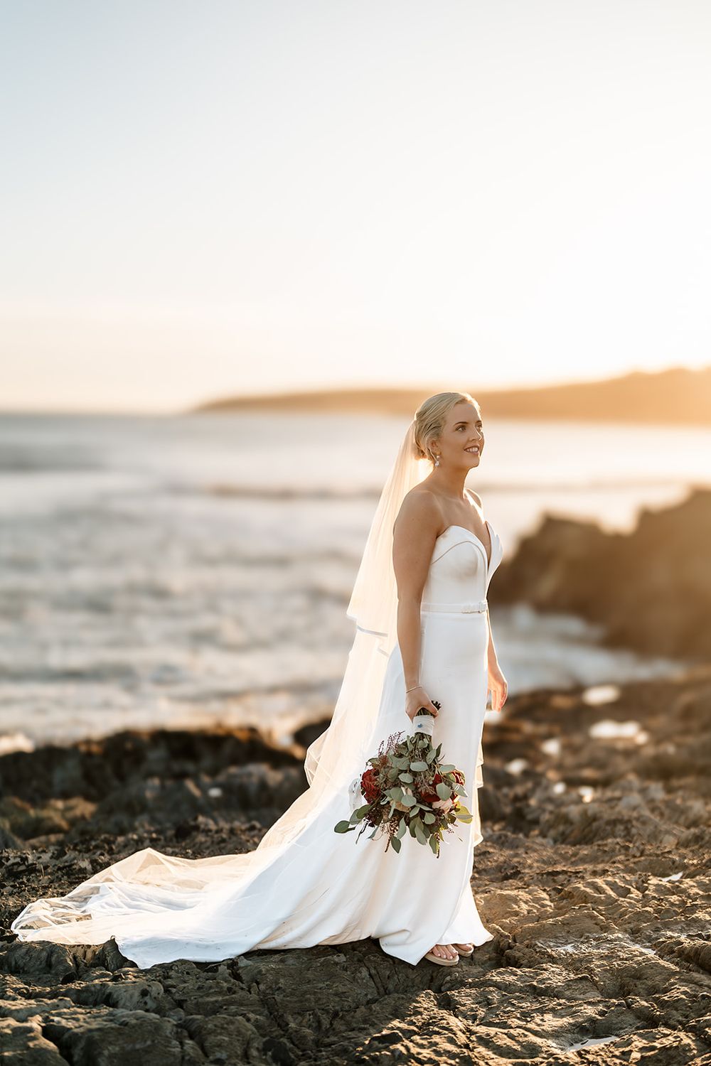Bride in flowing white dress poses on rocky coastline at sunset holding bouquet.
