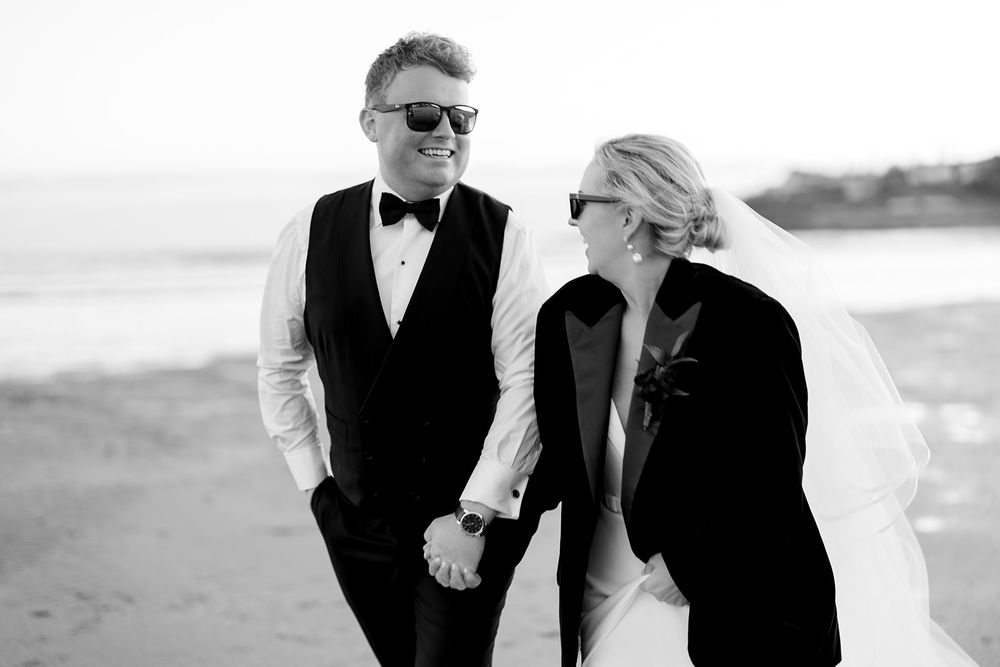 Black and white photo of couple in formal wear walking along beach at sunset.