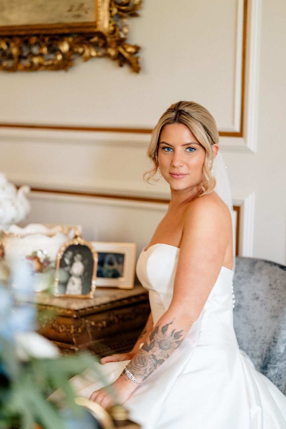 A bride in a white strapless wedding gown sits elegantly in an ornate room with gold-framed decor.