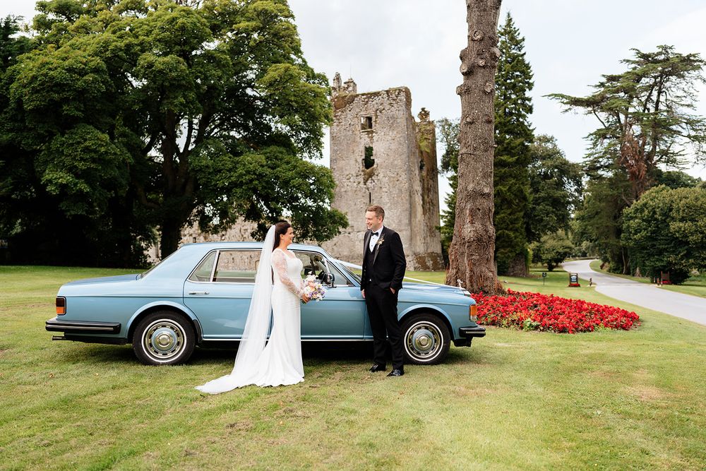 A vintage light blue car parked on a lawn in front of stone castle ruins and colorful flower gardens.