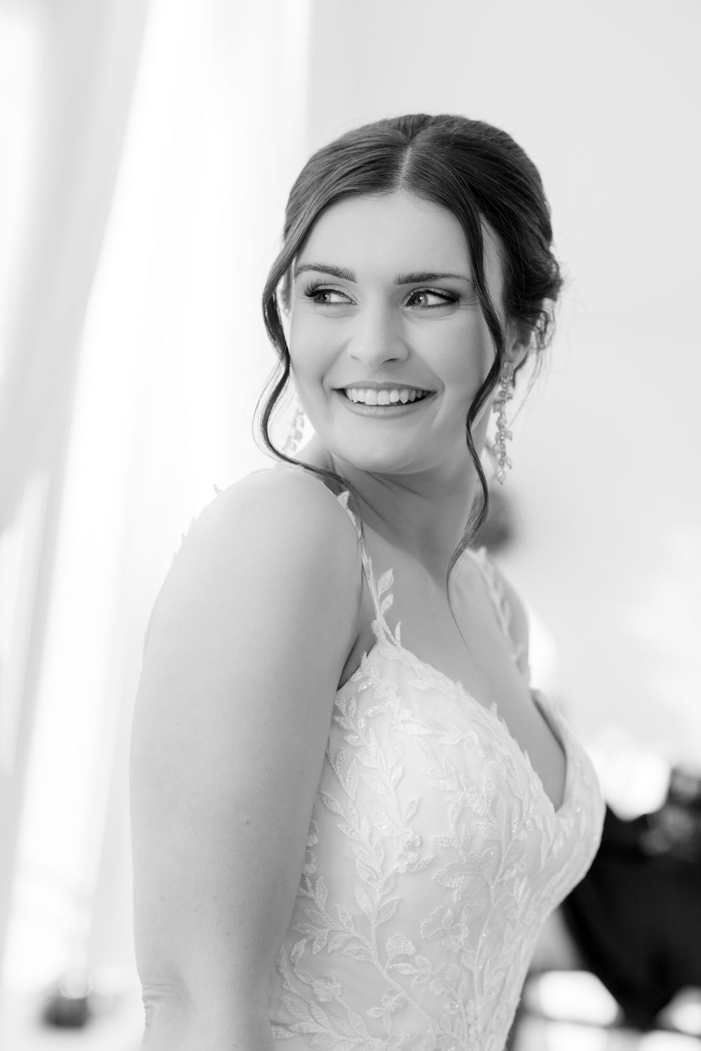 A black and white portrait of a smiling bride wearing a lace wedding dress with an elegant updo hairstyle.