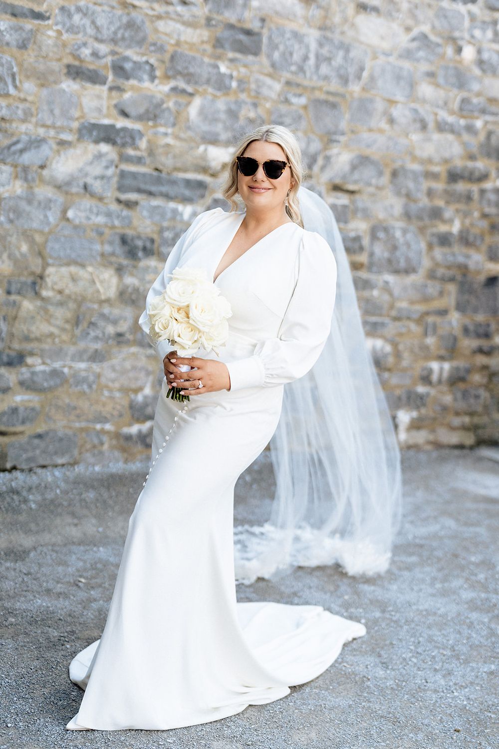 A bride in a sleek white long-sleeved wedding dress and sunglasses poses with bouquet against stone wall.