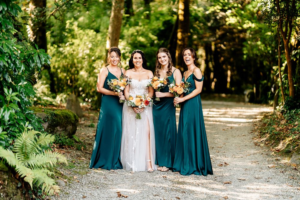Wedding party in elegant teal bridesmaid dresses standing on forest path surrounded by greenery.