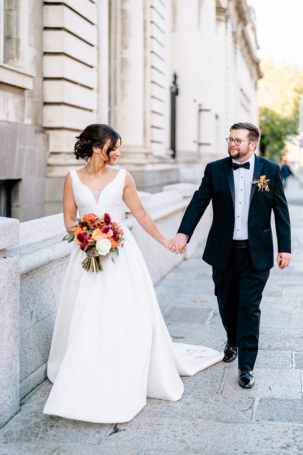 Elegant wedding couple walking hand in hand along stone building with bride holding autumn-colored bouquet.