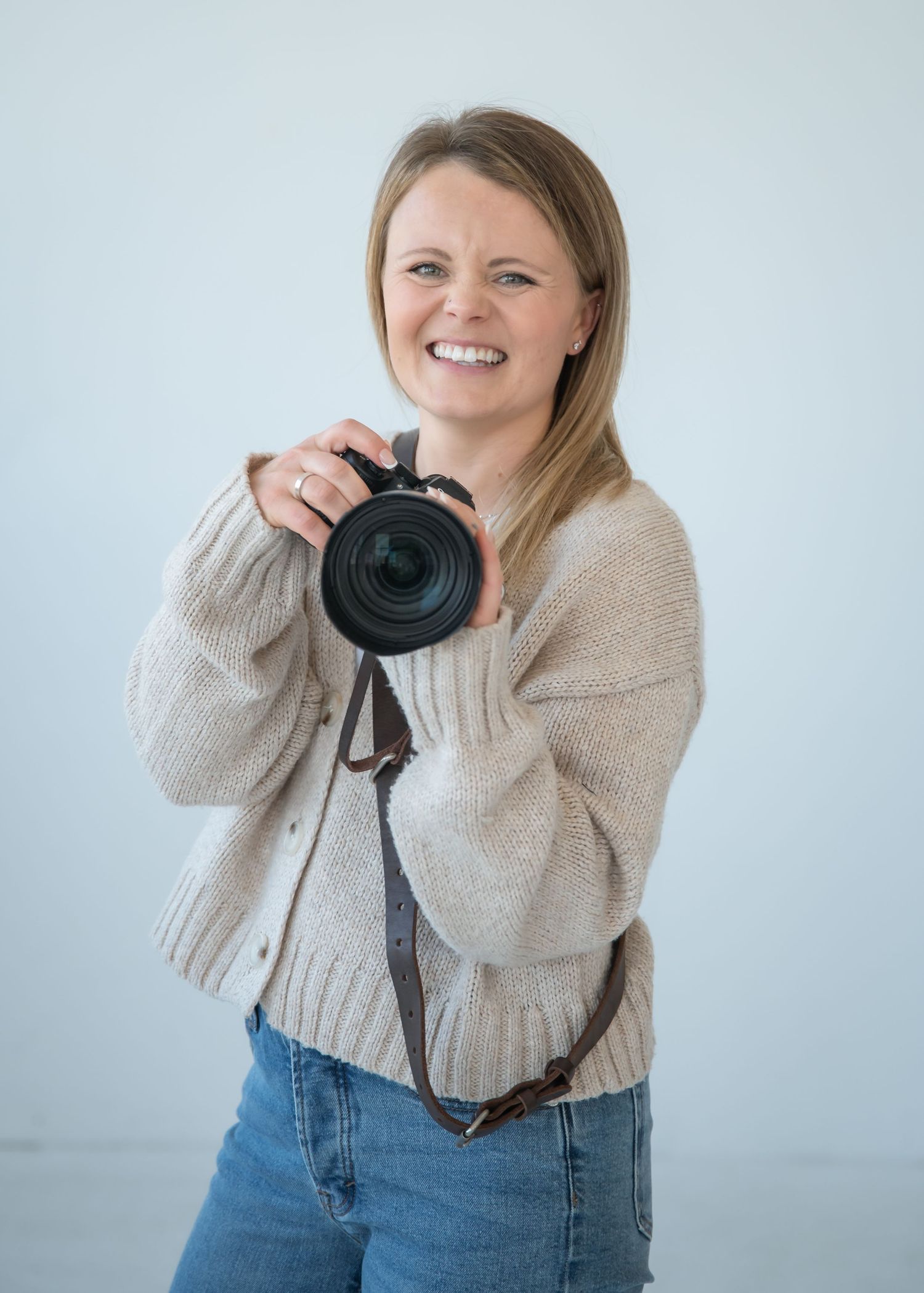 A photographer in a beige sweater and jeans holds a professional camera with a large lens against a light background.