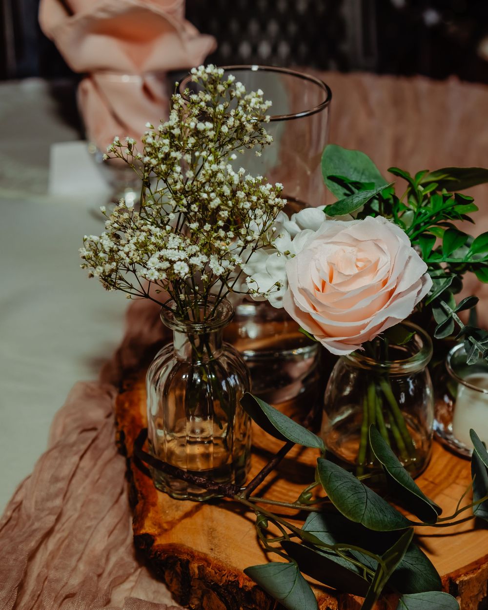 Delicate pink roses and baby's breath arranged in amber glass jars on rustic wooden slices create an elegant centerpiece.