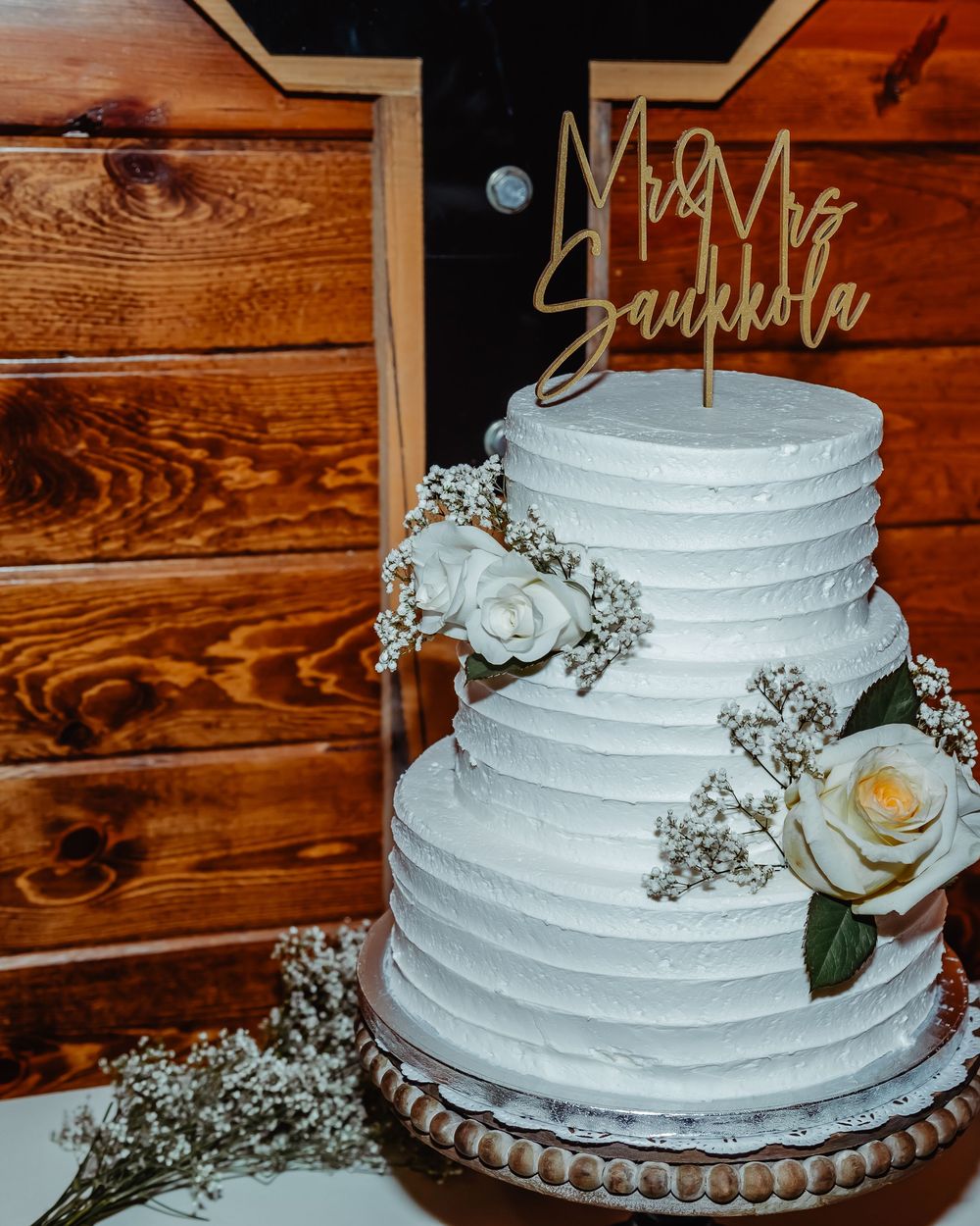 White buttercream wedding cake with textured icing, baby's breath flowers, and a single rose on a wooden backdrop.