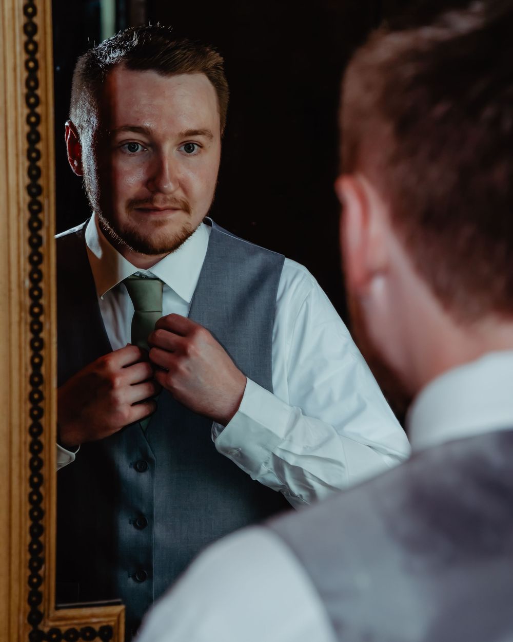 A groom adjusts his tie while looking in a framed mirror during wedding preparations.