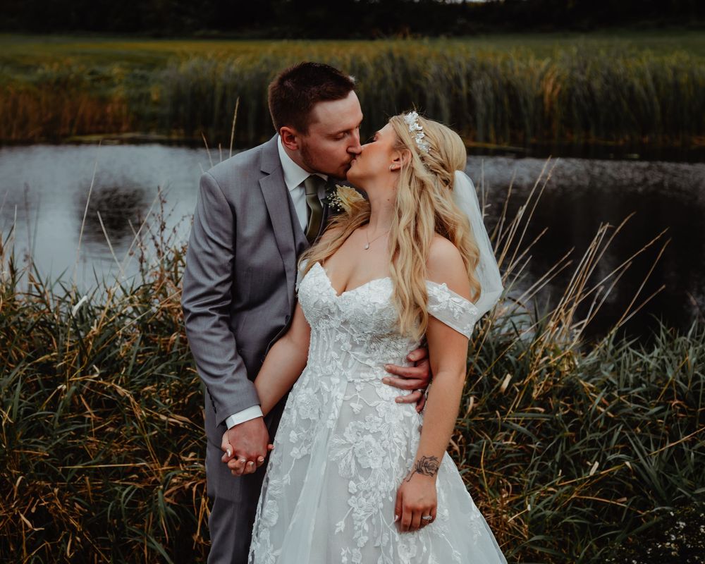 Newlyweds share an intimate kiss at sunset by a scenic pond surrounded by tall grass and reeds.
