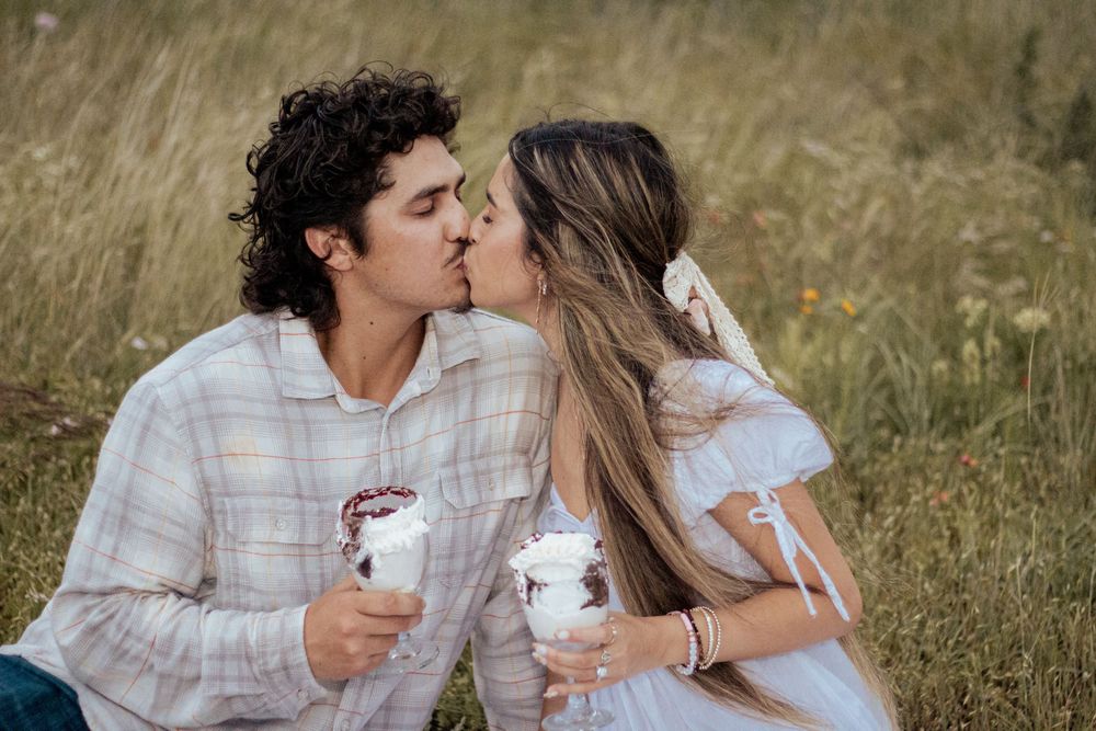 A romantic couple shares a kiss while holding coffee cups in a golden grass field during sunset.