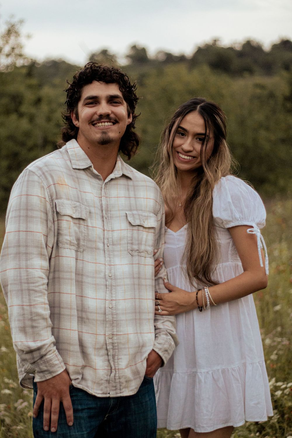 A couple stands together in a field at sunset, with the person in a plaid shirt and another in a white dress.