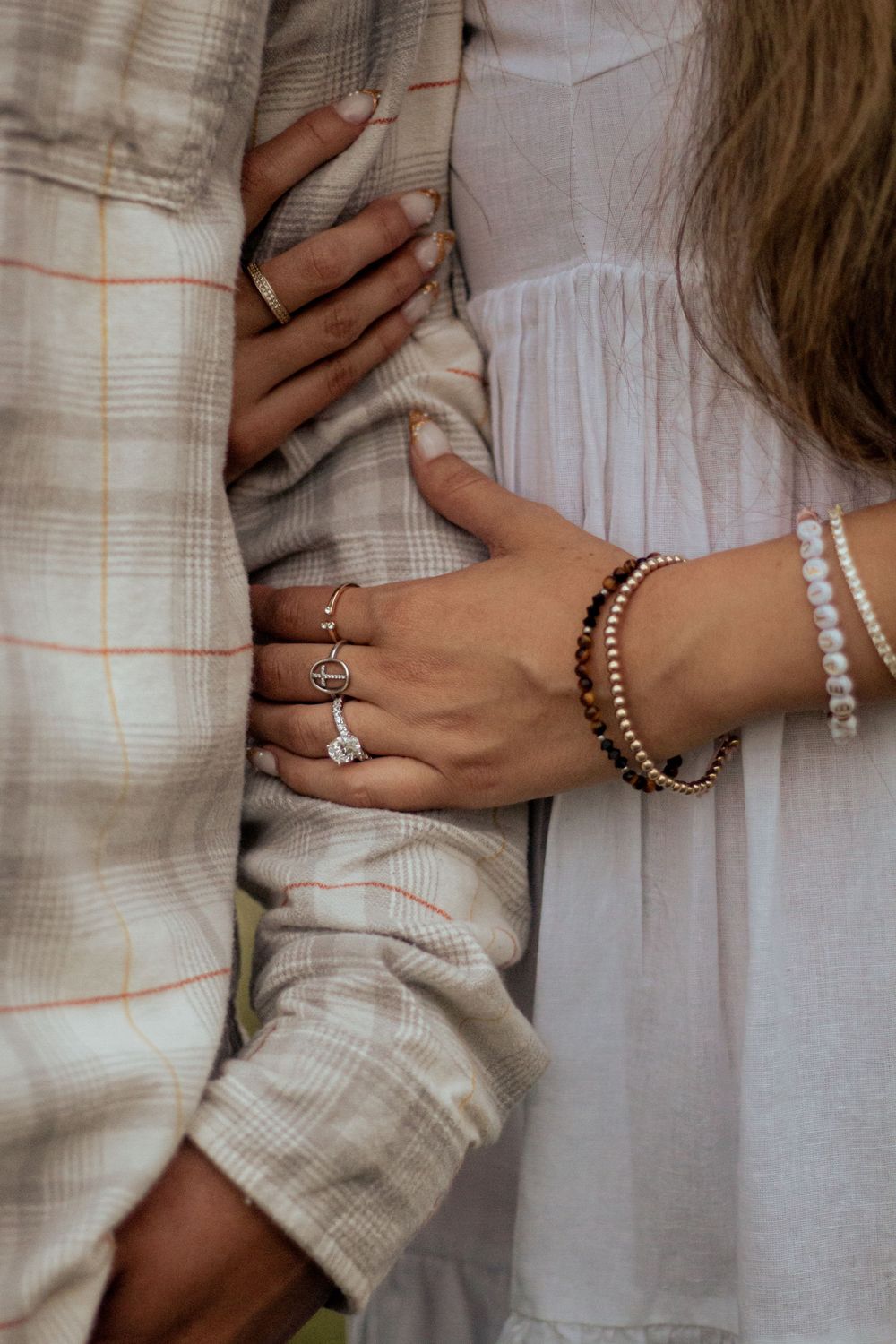 Close up of hands with multiple rings and bracelets against plaid shirt and white dress fabric.