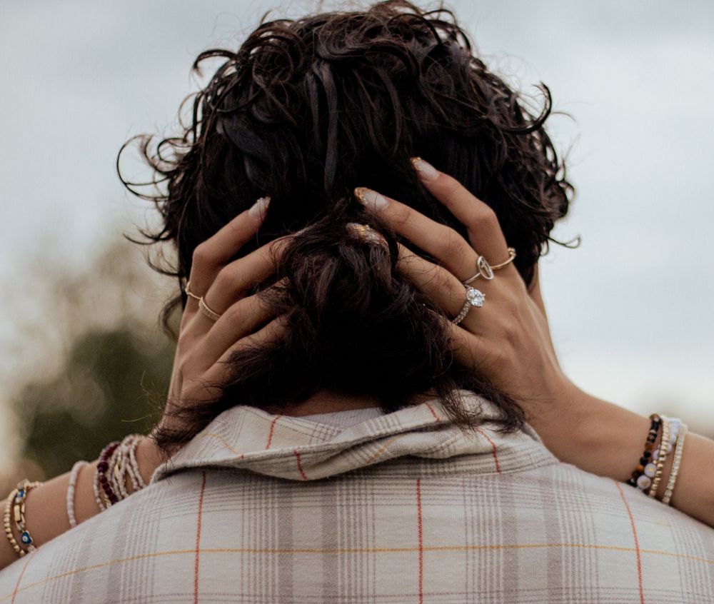 Back view of hands adorned with jewelry and rings running through dark curly hair against blurred outdoor background.