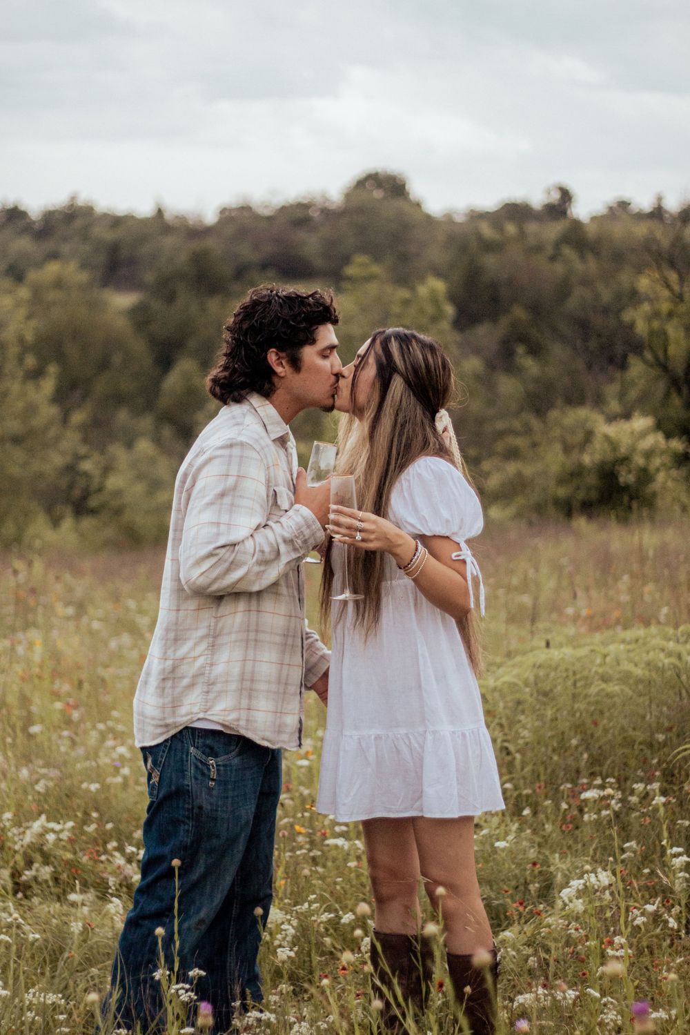 A couple shares a romantic moment while holding drinks in a wildflower meadow on an overcast day.