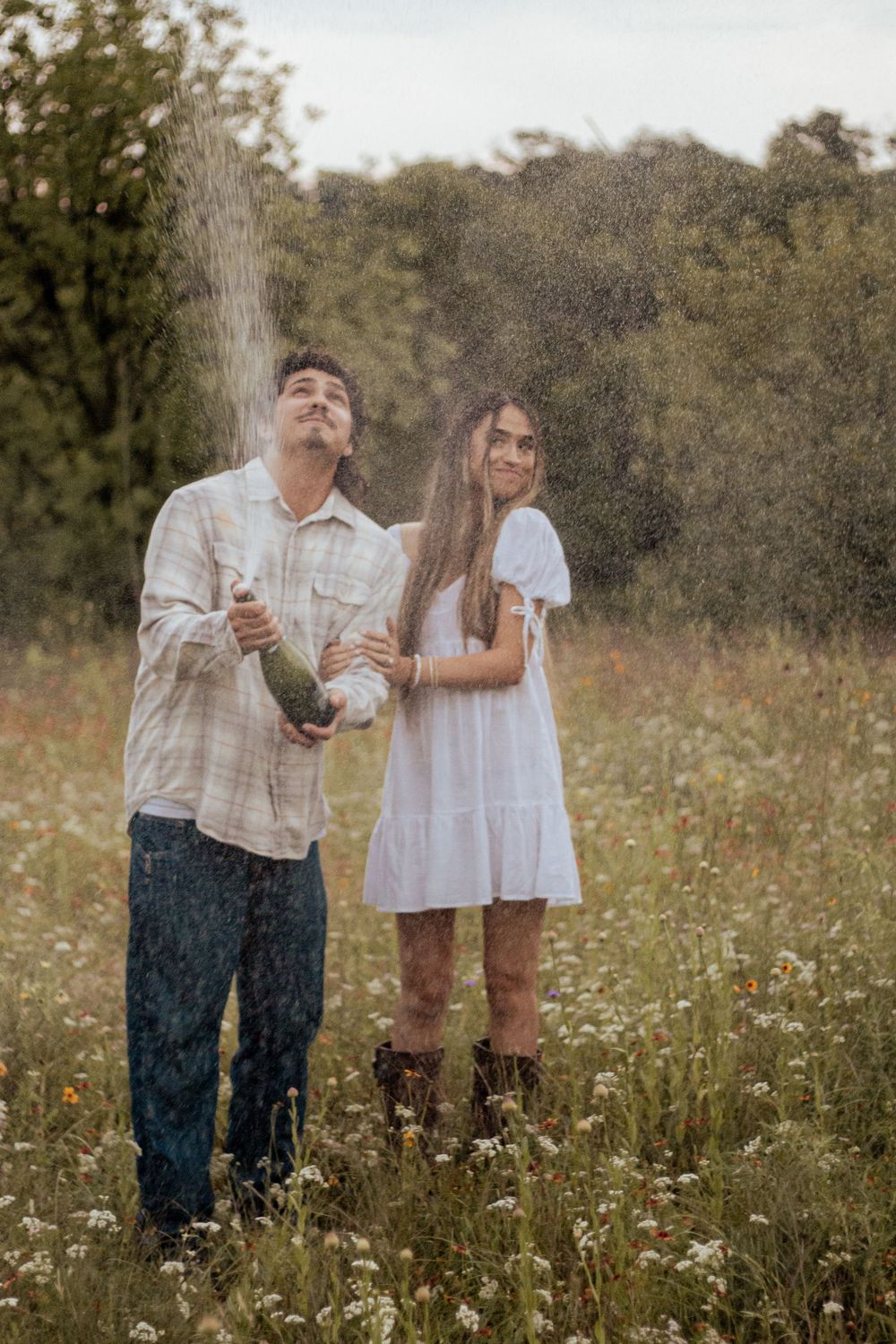 Two people stand in a meadow of wildflowers as water sprays through the air on a sunny afternoon in nature.