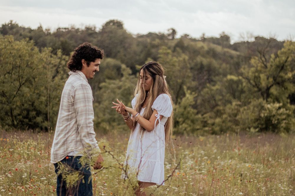 Two people in white outfits share a tender moment in a sunlit meadow filled with wildflowers.