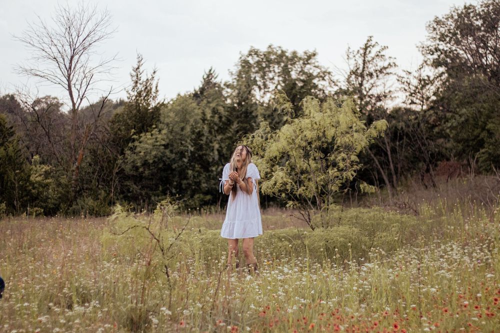 A figure in a light-colored dress stands among wildflowers in a serene meadow surrounded by trees.