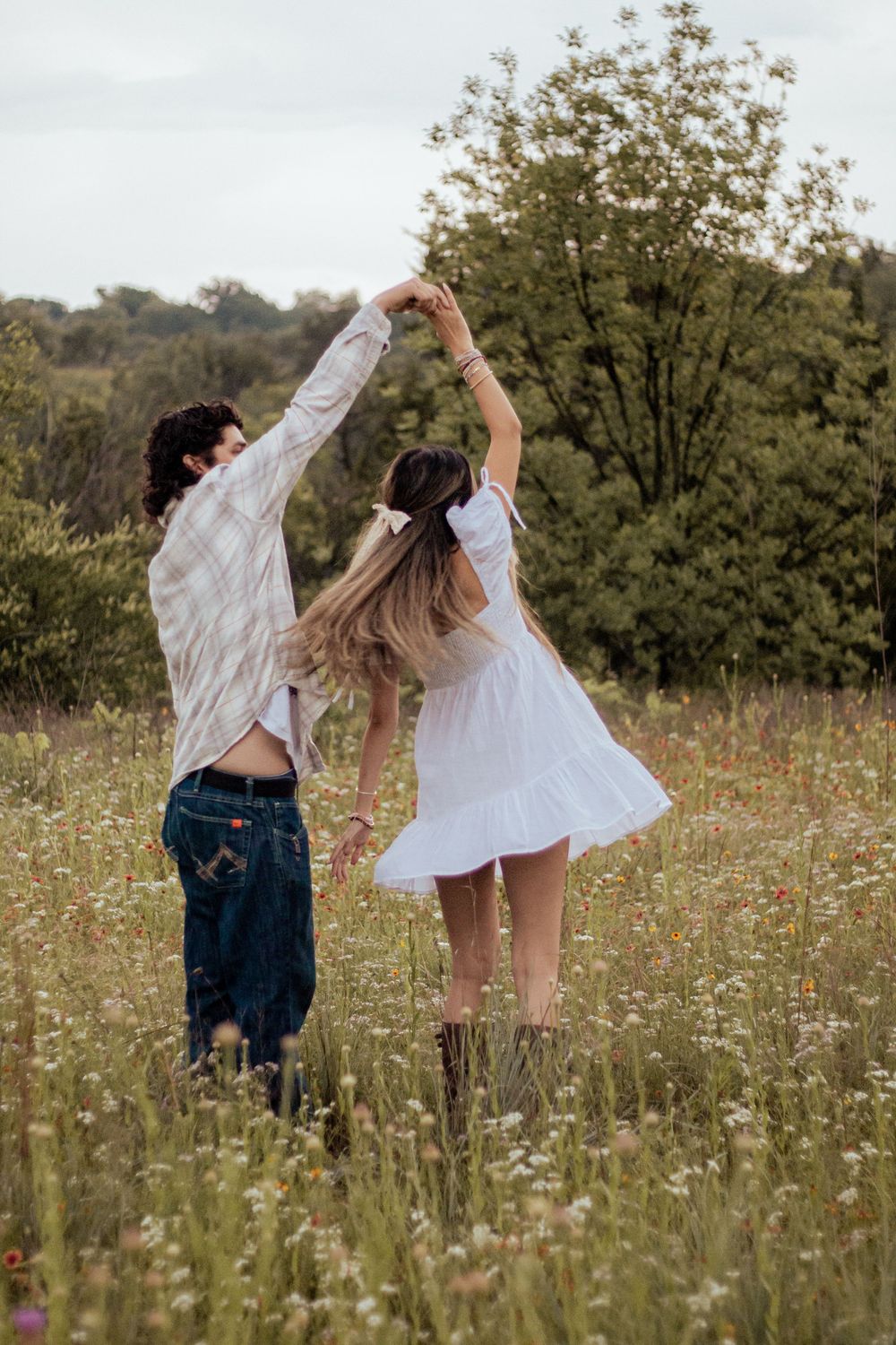 Couple dancing together in a wildflower meadow wearing white flowy outfits against green trees on a summer day.