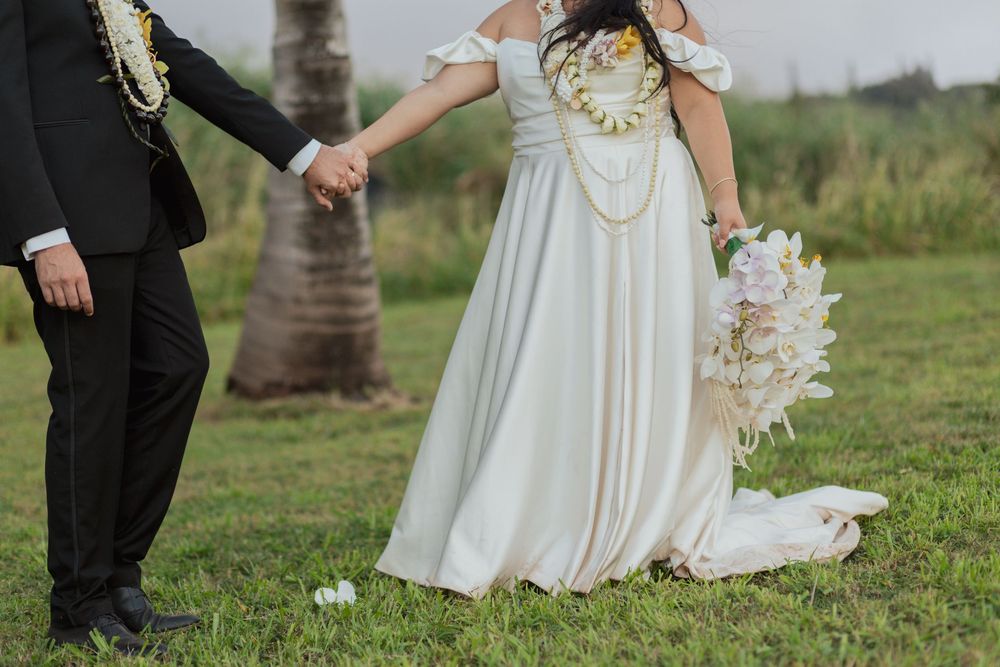 Close-up view of bride in flowing white dress and groom in dark suit holding hands outdoors on grass near a palm tree.