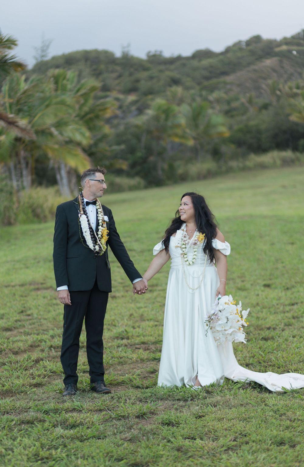 A couple in wedding attire hold hands on a grassy hillside with palm trees and mountains in the background.