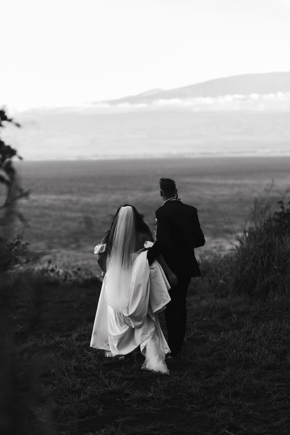 A bride and groom walk together on a hilltop overlooking a scenic landscape at sunset in black and white photography.