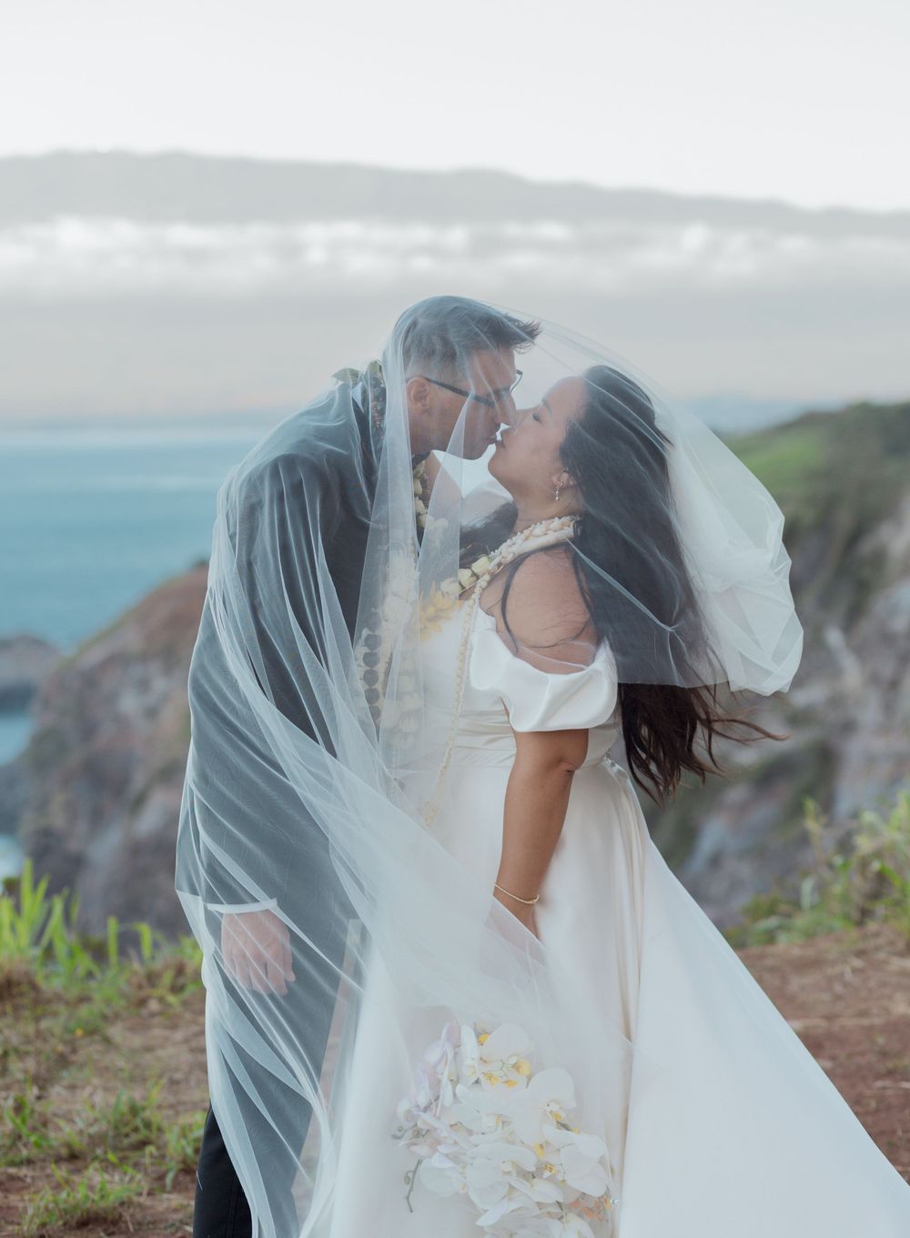 A couple embraces on a scenic coastal cliff with flowing veil and ocean views in the background.