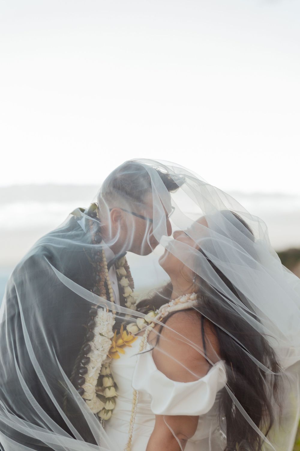 A romantic wedding couple shares an intimate moment under a flowing veil against a soft, dreamy background.
