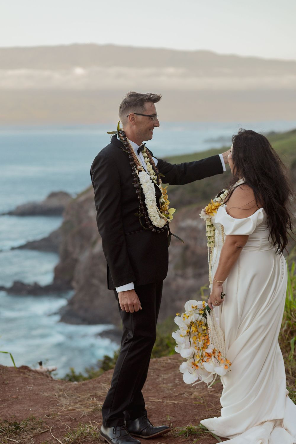 A couple in wedding attire embraces against a stunning ocean cliff backdrop at sunset.