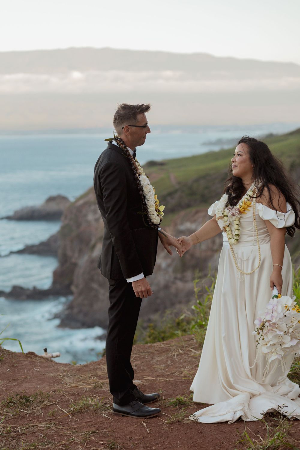 A couple wearing white and black attire exchange vows during an oceanside wedding ceremony on a scenic coastal cliff.